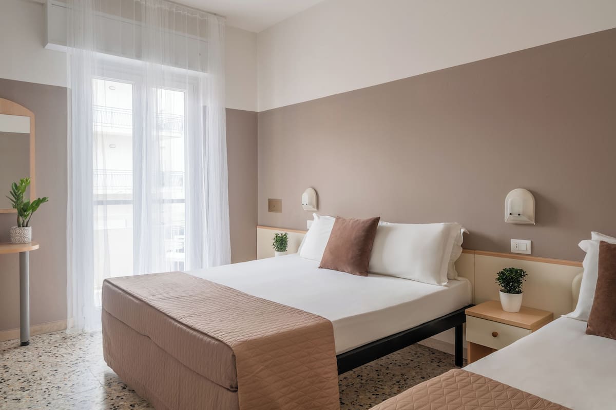 A hotel room with a large bed, bedside tables with plants, a mirror, and a window with sheer curtains.