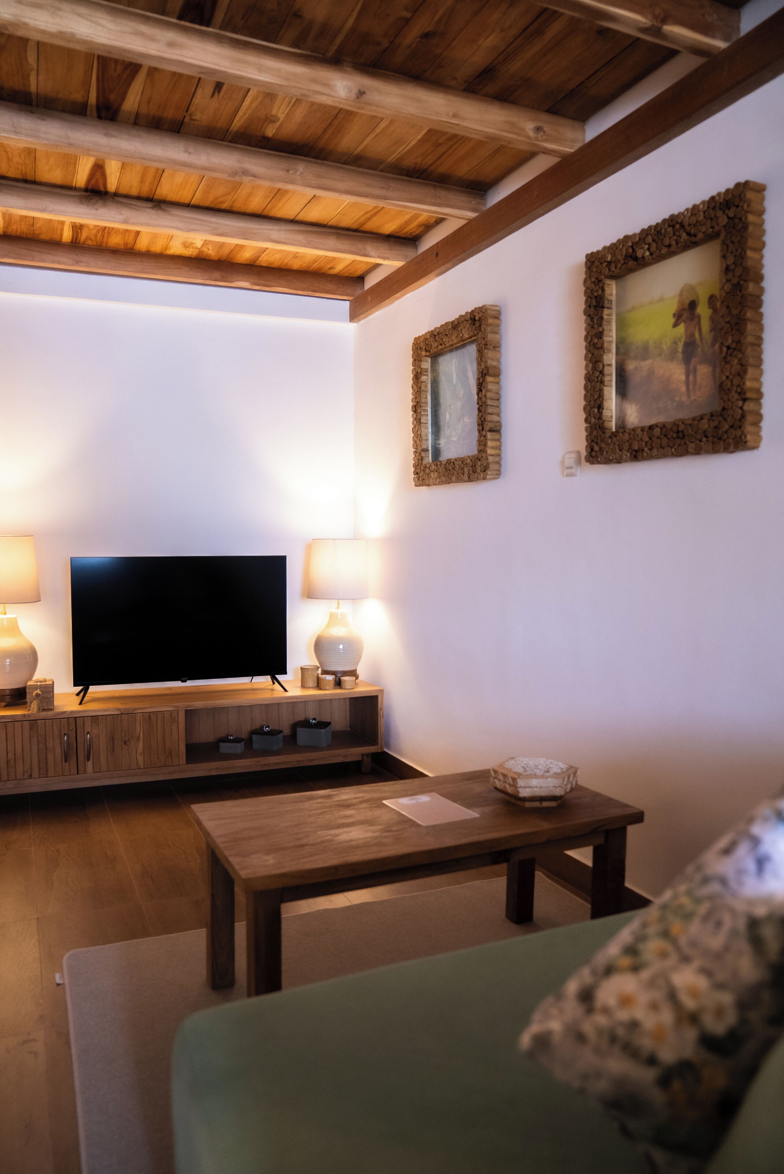 A cozy living room with a wooden ceiling, a flat-screen TV on a wooden stand, two wall-mounted lamps, and a green sofa.
