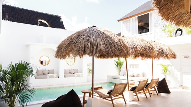 Un espace aménagé au bord de la piscine, avec des chaises longues, un parasol en chaume et un bâtiment blanc doté d’un toit en tuiles.