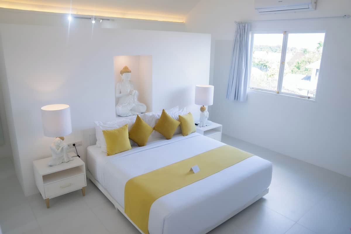 Een moderne slaapkamer met een groot bed, witte wanden, een Boeddhabeeld en een raam met uitzicht.