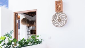 Un coin de pièce avec un mur blanc, une étagère en bois, deux tabourets en osier, une décoration murale tissée et une fenêtre donnant sur une autre pièce.