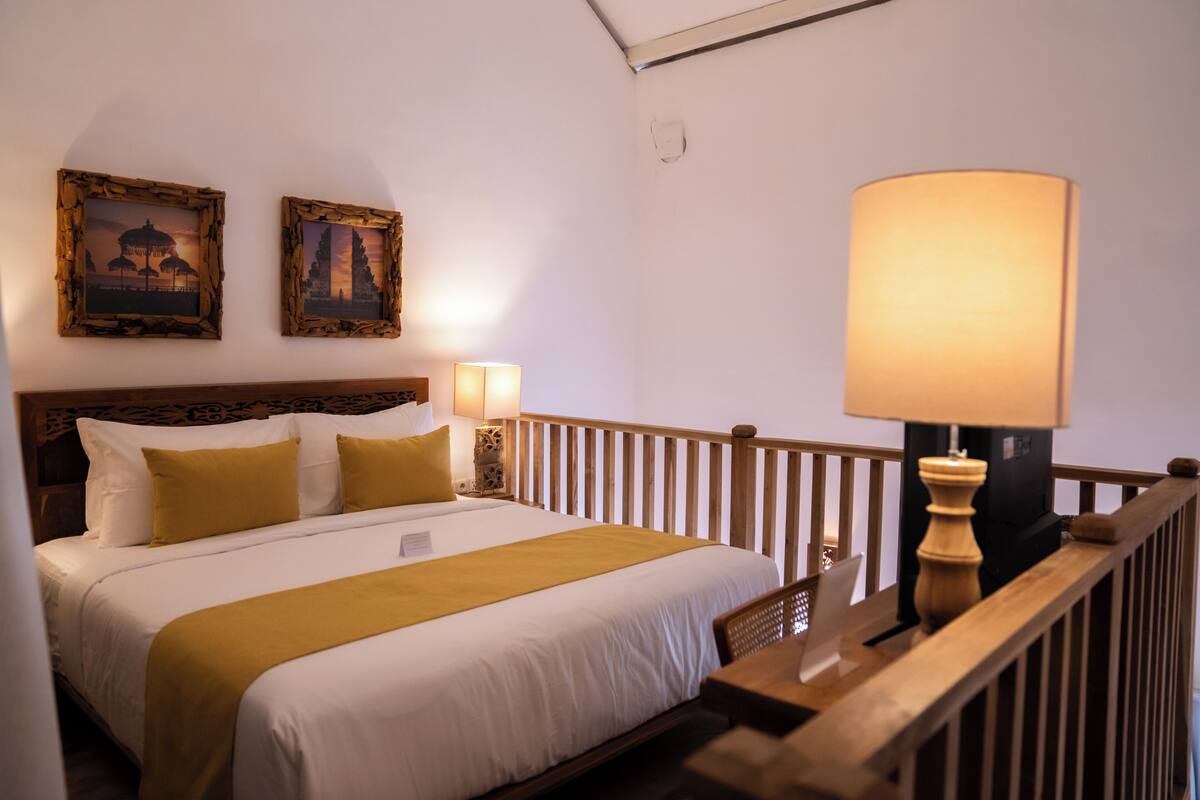 Een netjes opgemaakt bed met een houten hoofdbord, twee bedlampen en twee afbeeldingen in lijsten aan de muur.