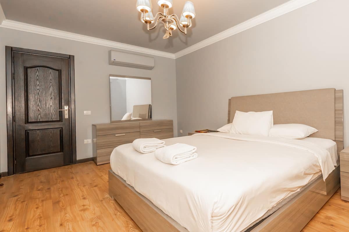 A modern bedroom with a wooden floor, a large bed with white linens, a bedside table, a nightstand, a dresser, and a chandelier.