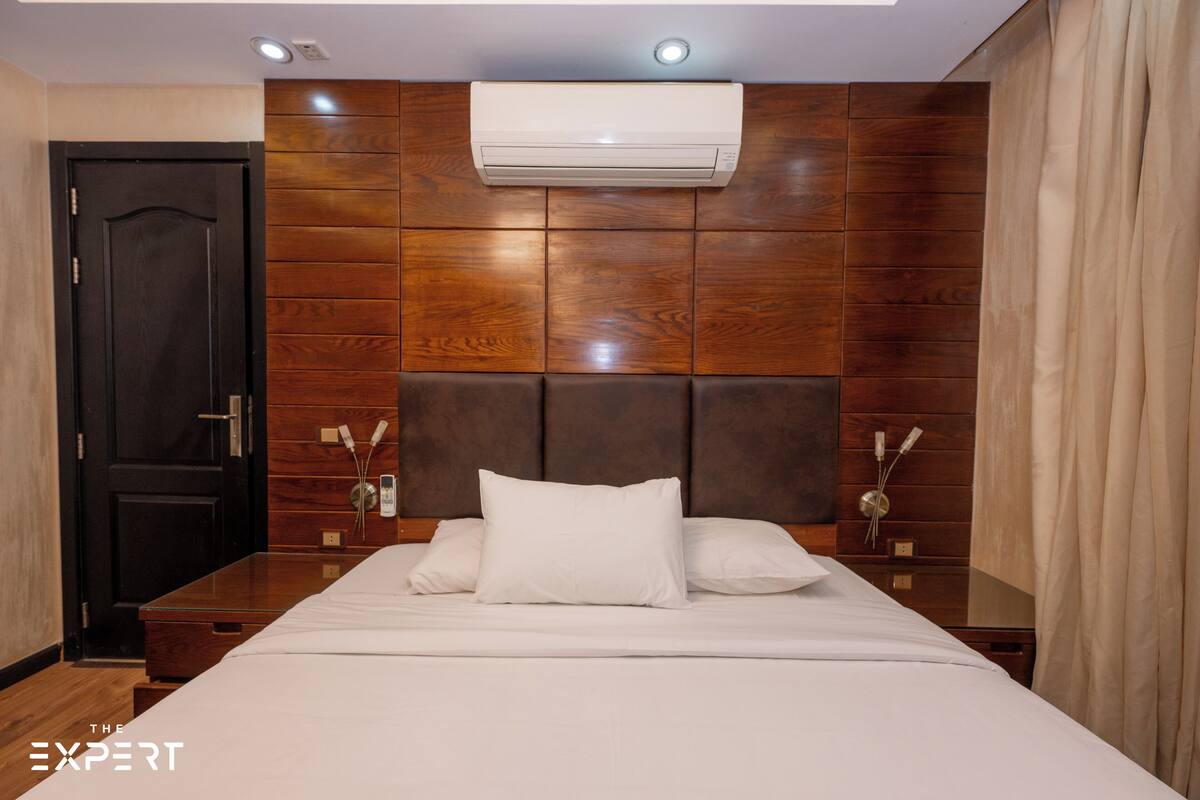A hotel room with a wooden headboard, a bed with white linens, a dark door, and an air conditioning unit mounted on the wall.