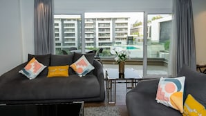 A modern living room with a grey sofa set, a coffee table, and a view of a swimming pool and apartment buildings through large windows. - The Expert (Cairo)