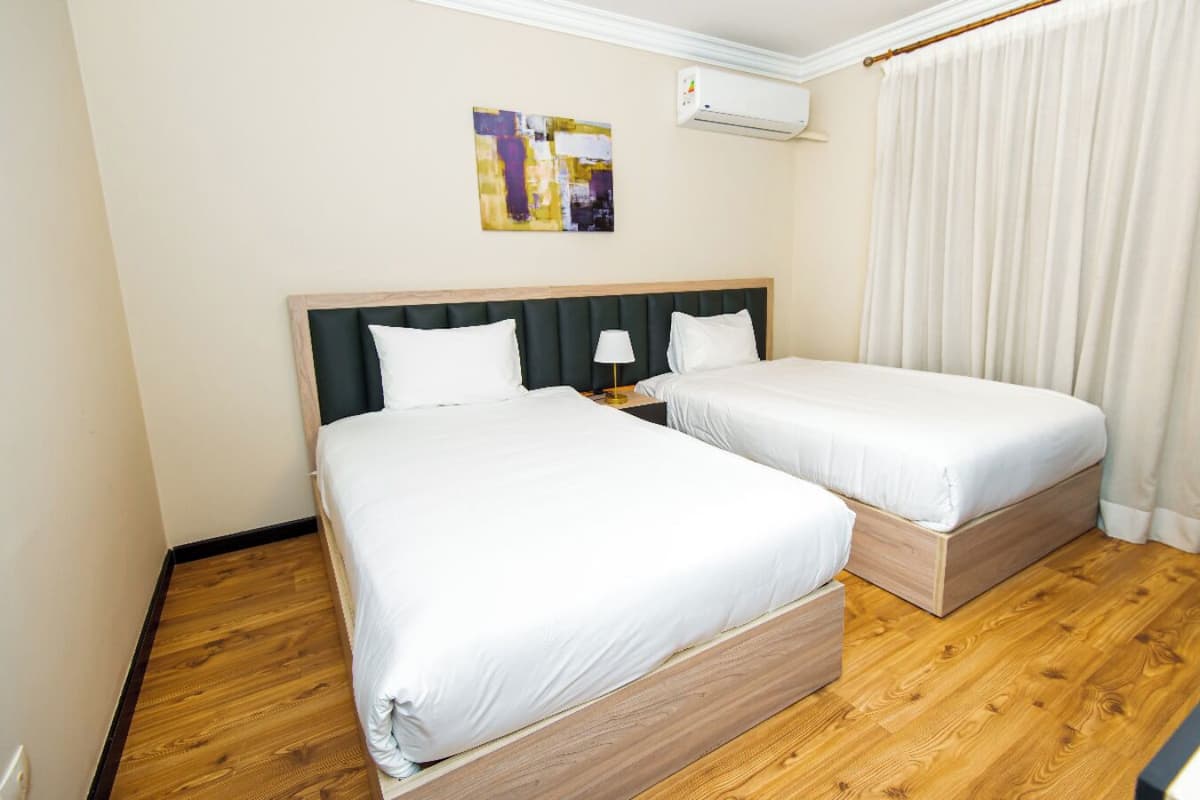 A hotel room with two beds, wooden flooring, a ceiling fan, an air conditioning unit, and a painting on the wall.