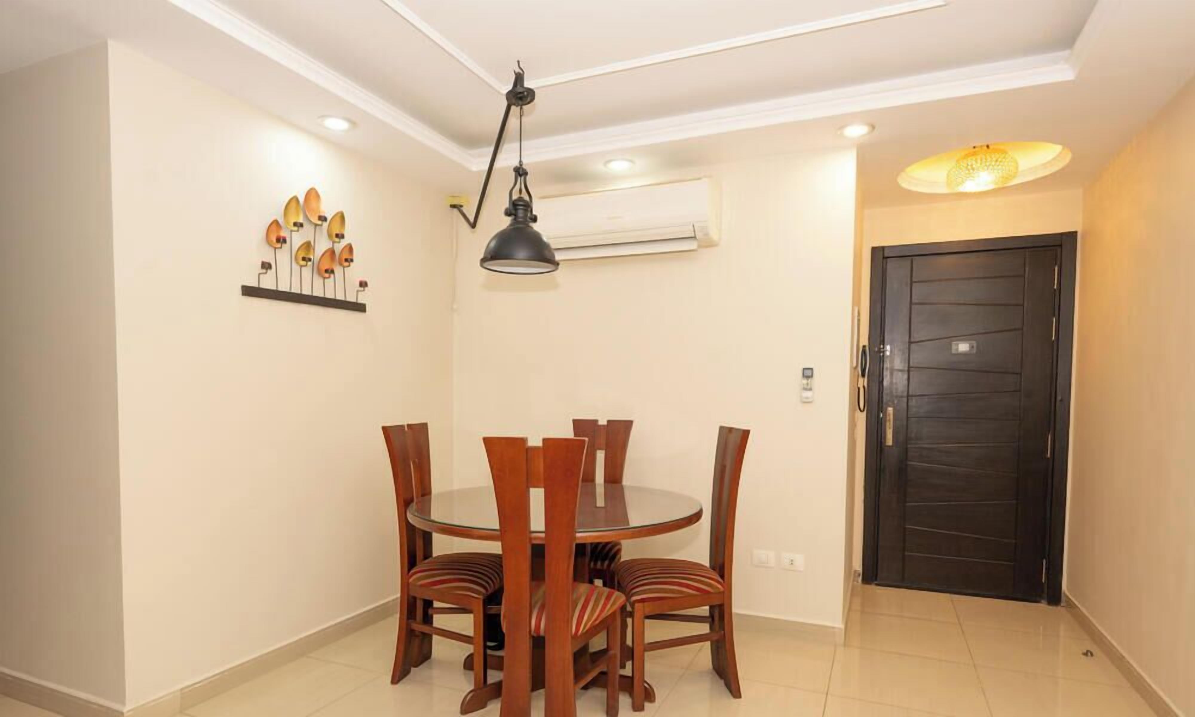 A dining area with a round table and six chairs, a pendant light, an air conditioning unit, and a dark door.