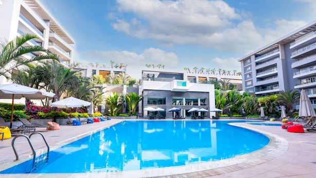 A swimming pool with lounge chairs and umbrellas, surrounded by greenery and modern buildings. - The Expert (Cairo)