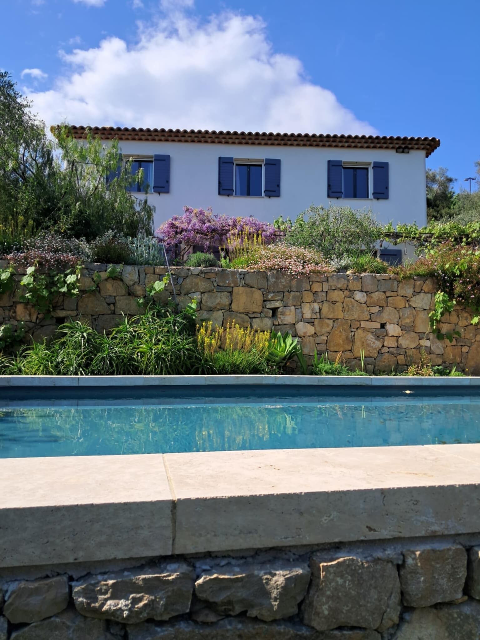 A white building with blue shutters, a stone wall, and a swimming pool.