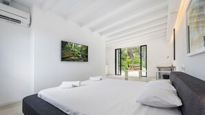 A modern bedroom with a large bed, a flat-screen TV, and a view of a garden through a sliding door. - My Rental Homes - Can Cabana with private pool, air conditioning and wifi (Pollença)