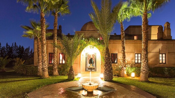 A well-lit courtyard with a fountain, palm trees, and a building with arched windows and a dome. - My Rental Homes - Dar Maril with pool, tennis court and garden (Oulad Jellal)