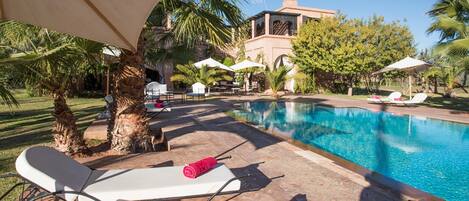 Zona de piscina con sillas de descanso y sombrillas, rodeada de palmeras y un edificio con torre.
