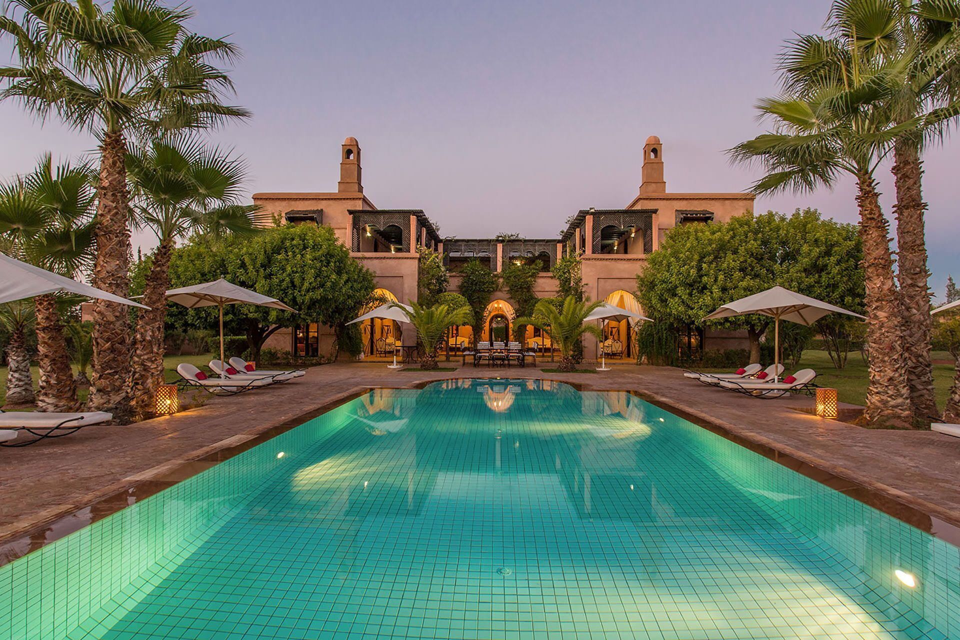 A swimming pool with lounge chairs and umbrellas, surrounded by palm trees and a building with arched windows.
