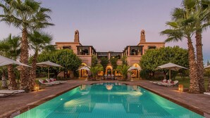 A swimming pool with lounge chairs and umbrellas, surrounded by palm trees and a building with arched windows. - My Rental Homes - Dar Maril with pool, tennis court and garden (Oulad Jellal)
