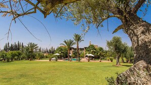 A well-maintained lawn with a pool, palm trees, and a building with a clock tower in the background. - My Rental Homes - Dar Maril with pool, tennis court and garden (Oulad Jellal)