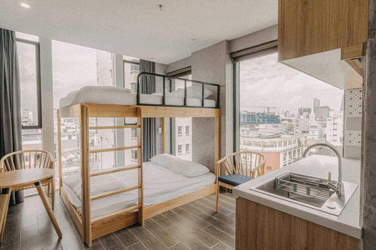 Una cama literera con escritorio y silla, una pequeña cocina con lavamanos, y ventanas amplias con vistas a la ciudad.