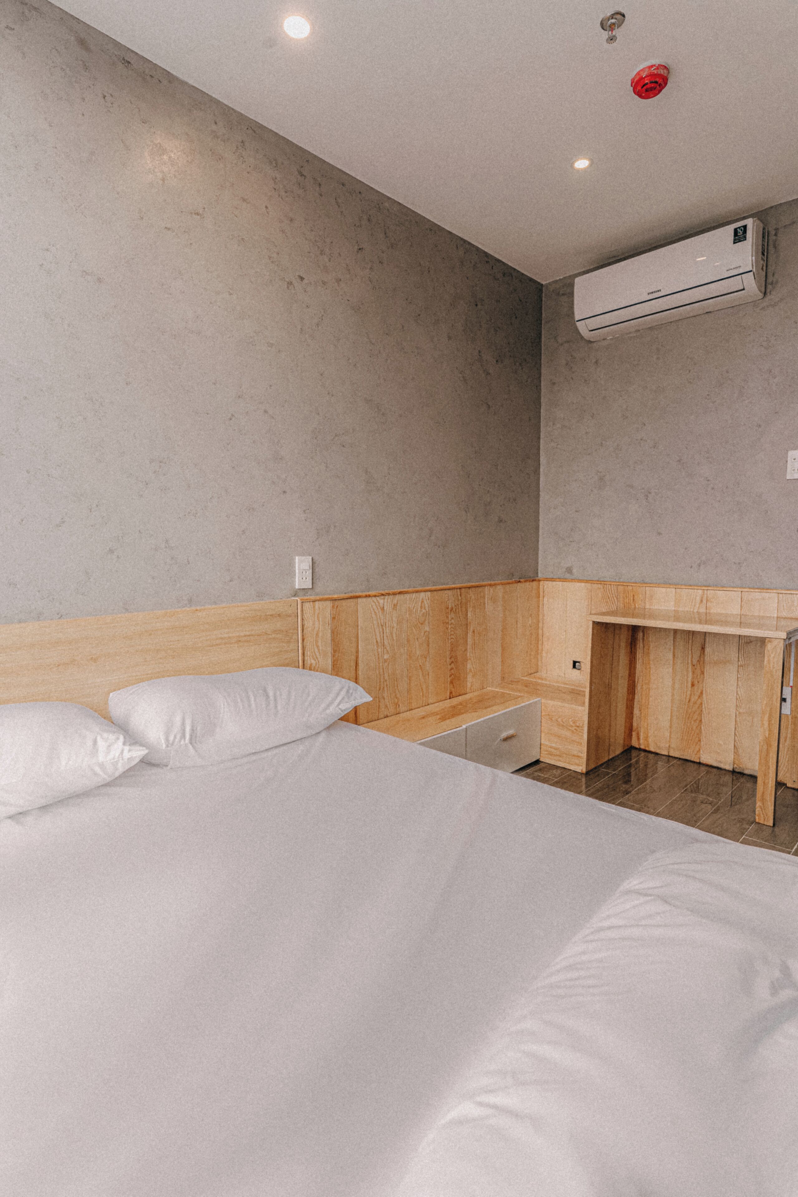 A modern hotel room with a wooden headboard, a single bed with white linens, a small wooden desk, and a wall-mounted air conditioning unit.