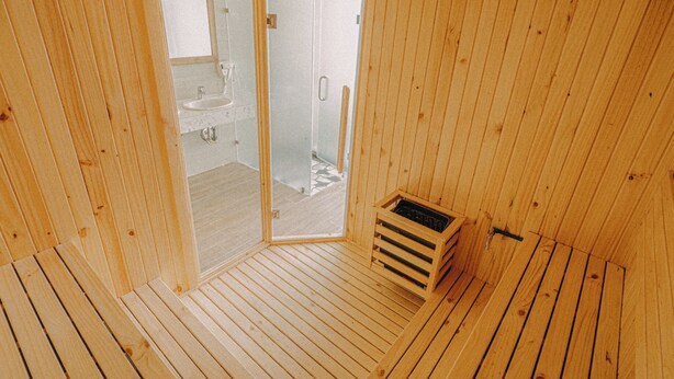 A wooden cabin interior with a wooden floor, a small wooden bench, and a glass door leading to a bathroom with a sink and shower. - SNite Hotel & Dorm-Danang Dragon Bridge (Da Nang)
