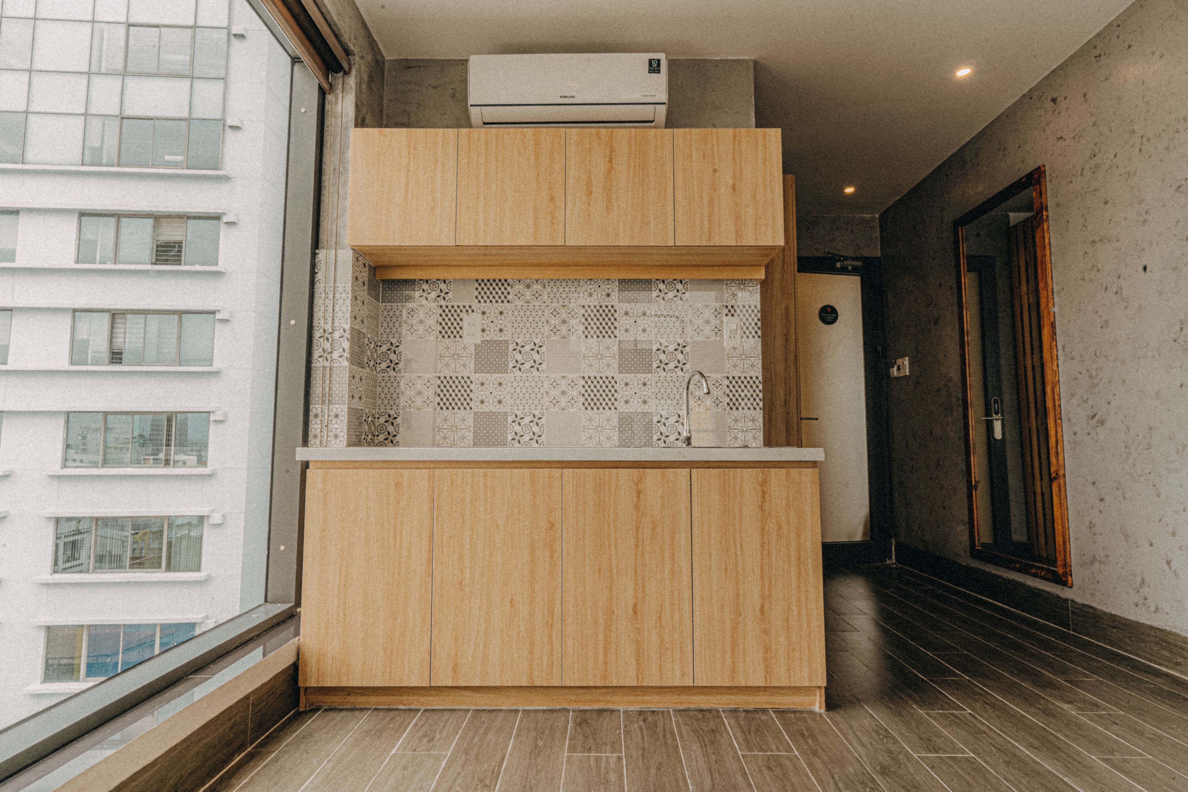 A compact kitchen with a built-in air conditioner, a patterned backsplash, and a view of a building outside.