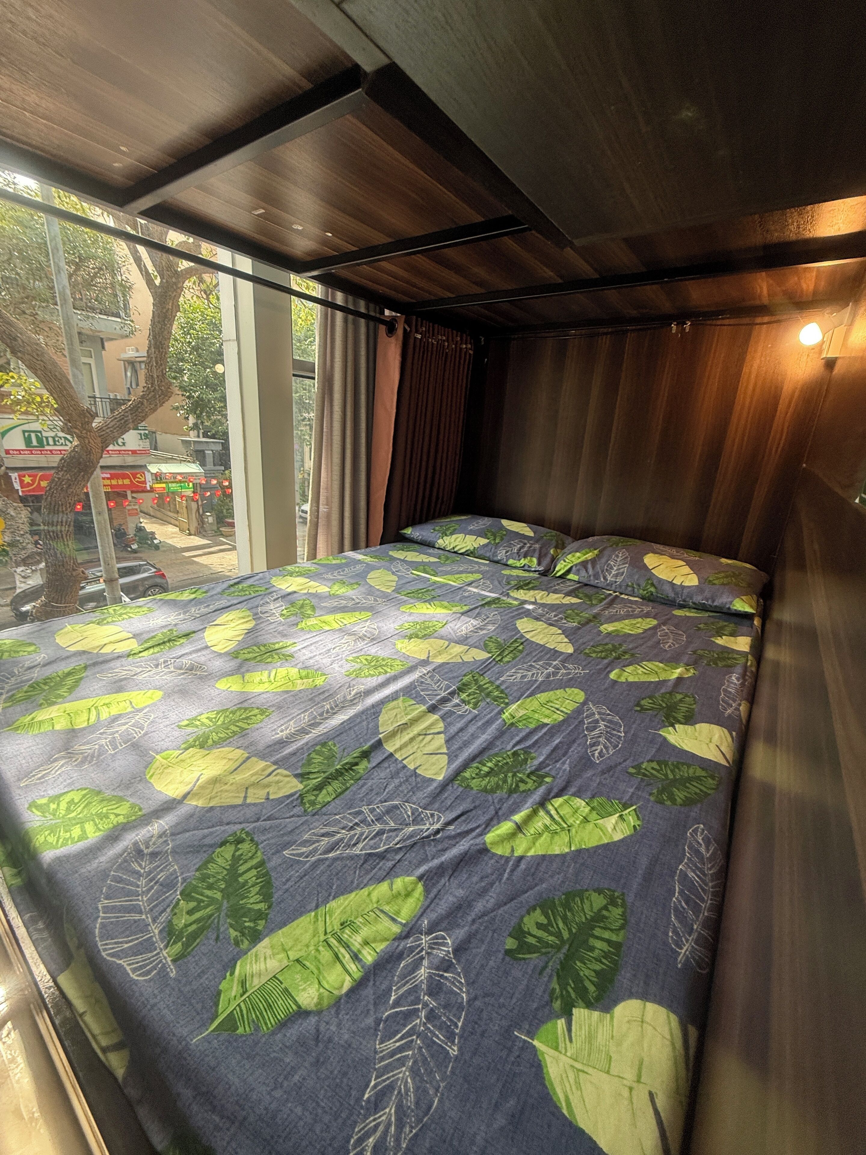 A compact room with a bed featuring a leaf-patterned bedspread, a window with curtains, and a view of the street outside.
