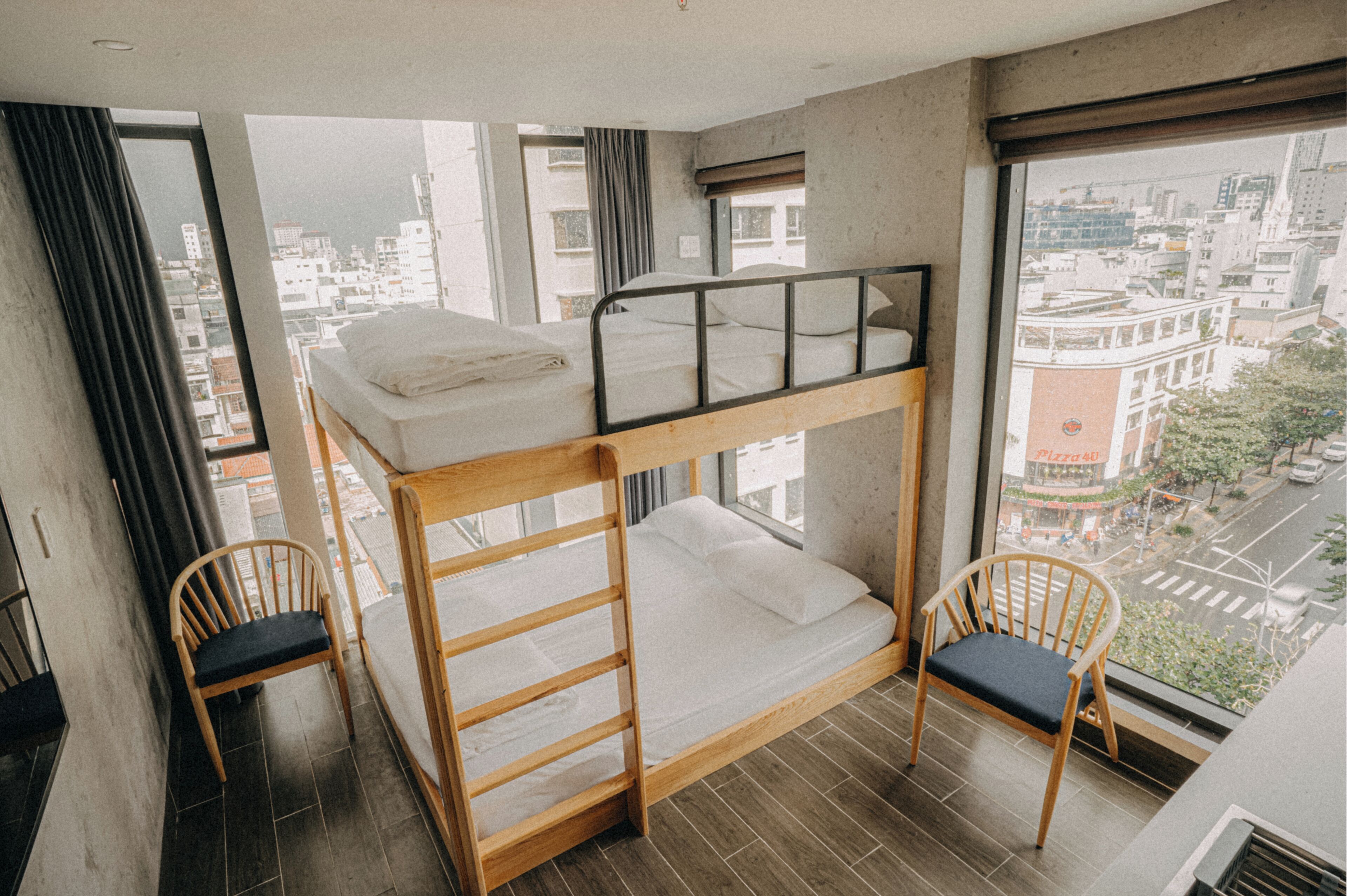 A bunk bed room with a city view, two wooden chairs, and a large window.
