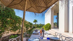Zona d'esmorzar a l'aire lliure amb una taula preparada per a dues persones, un parasol trenat i vista del paisatge circumdant.