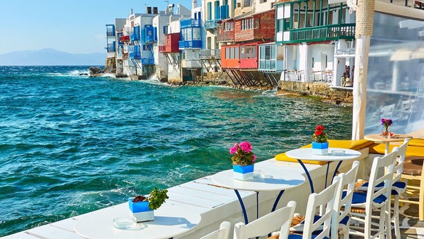 Un restaurante frente al mar con terraza al aire libre, mesas blancas y sillas azules, y edificios coloridos al fondo.