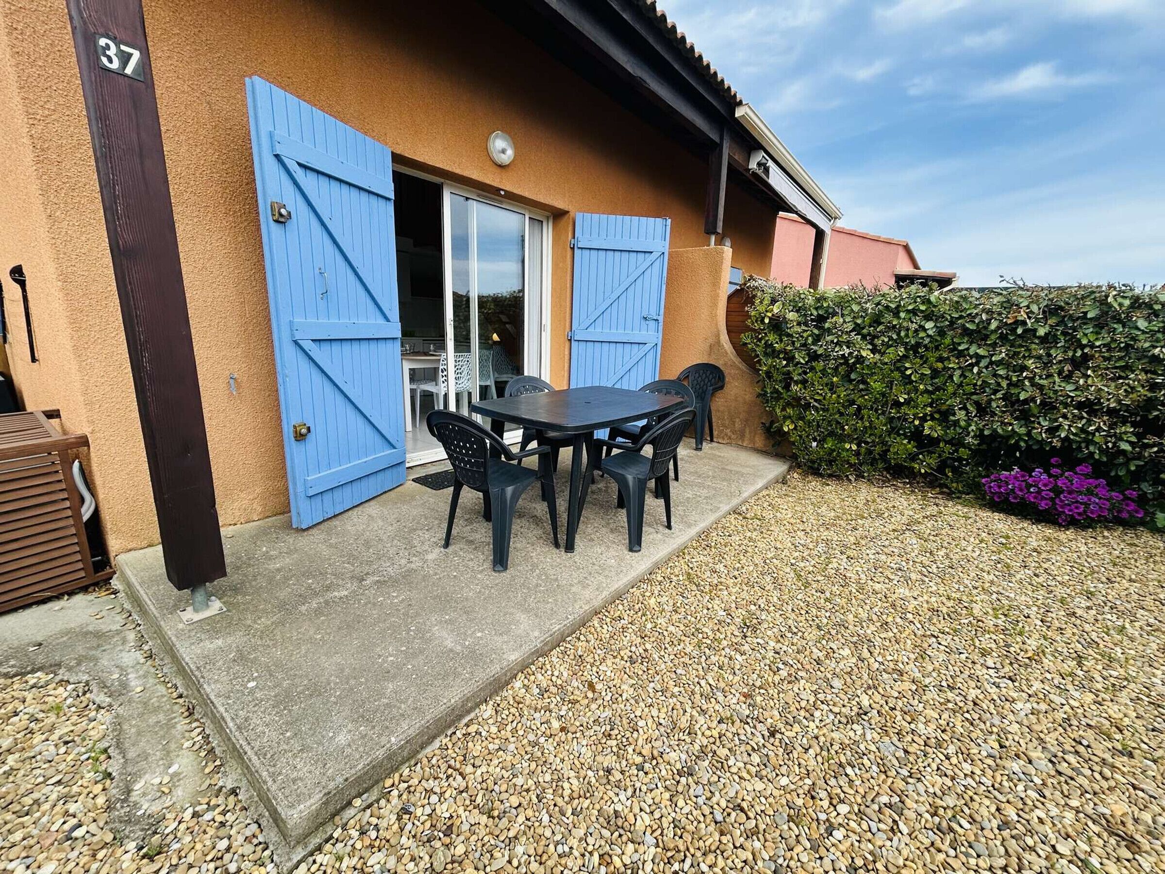 A building with a number '37' sign, a blue door, a patio with a table and chairs, and a hedge with purple flowers.