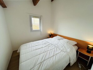 A single bed with a white duvet, a nightstand with a lamp, a window with curtains, and a wooden ceiling beam. - 3-Room Pavilion, Terrace, Narbonne Beach (Narbonne)