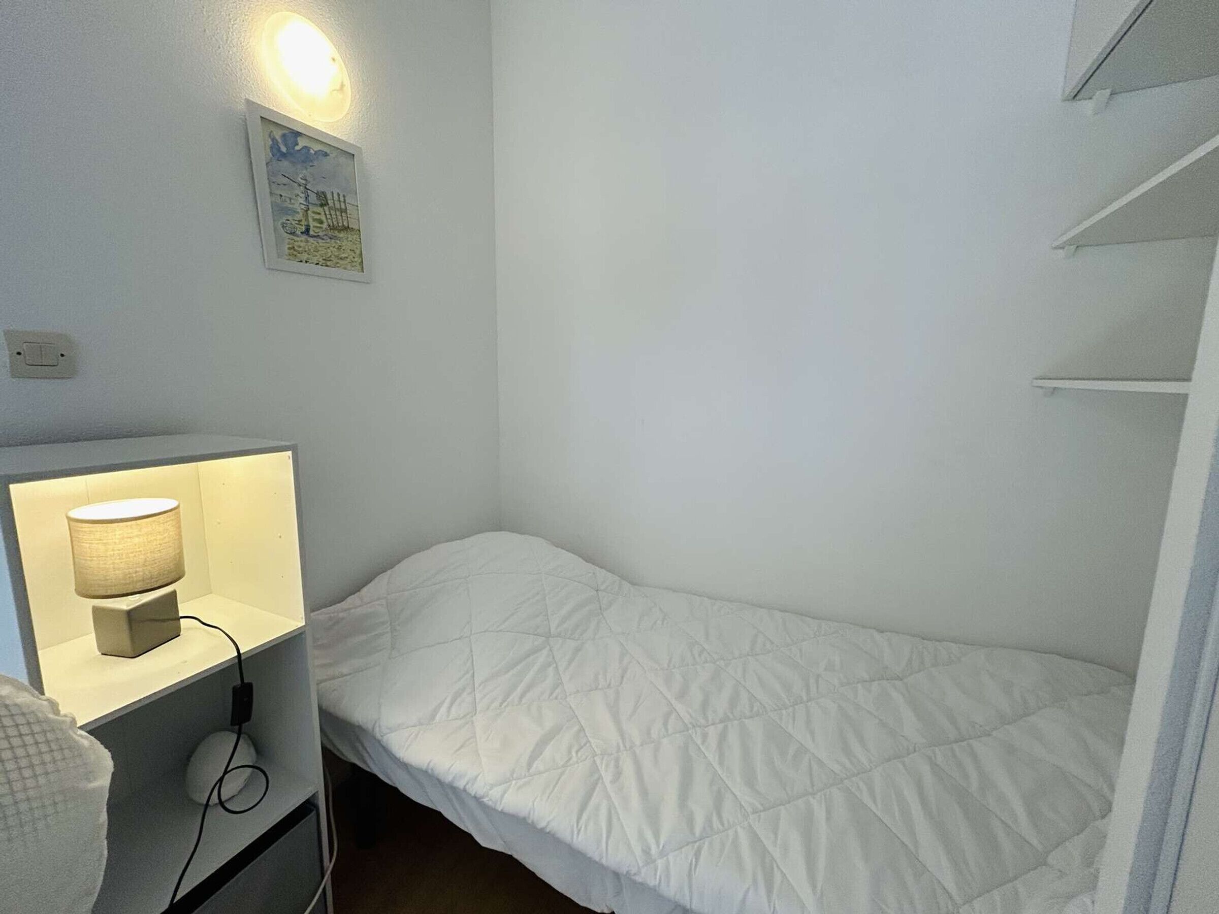 A small, single-bed room with a bedside lamp, a power outlet, and a wall-mounted light fixture.