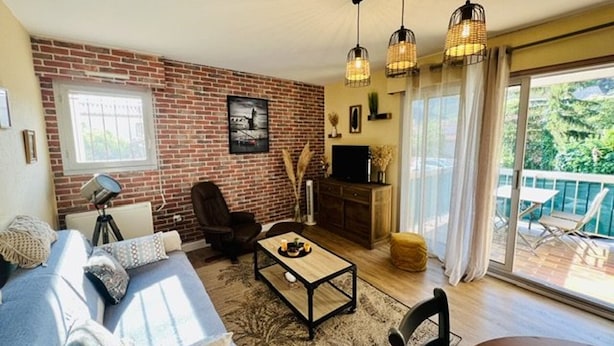 A living room with a brick wall, a sofa, a coffee table, a TV, and a balcony with outdoor furniture.