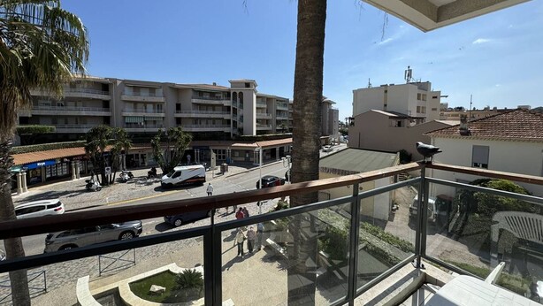 A balcony with a clear view of a street, palm trees, and a multi-story building with a tiled roof.
