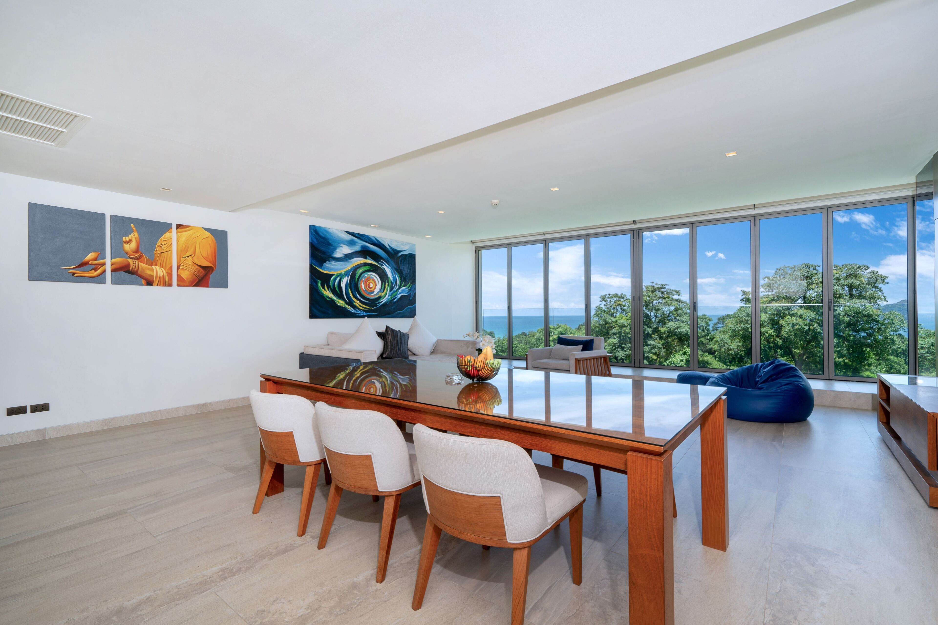 A spacious modern living room with a glass dining table, white chairs, and a large window offering a view of the outdoors.