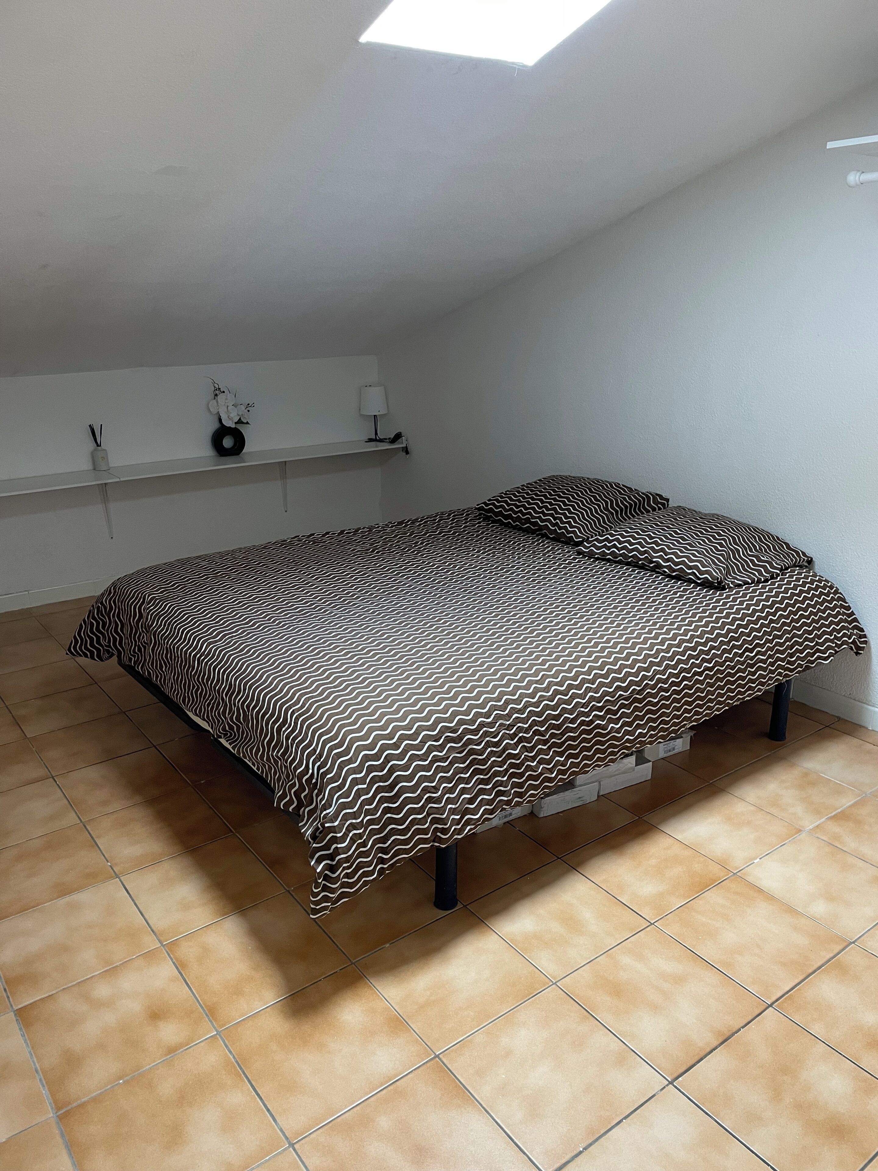 A bedroom with a bed, a skylight, a shelf with decorative items, and a tiled floor.