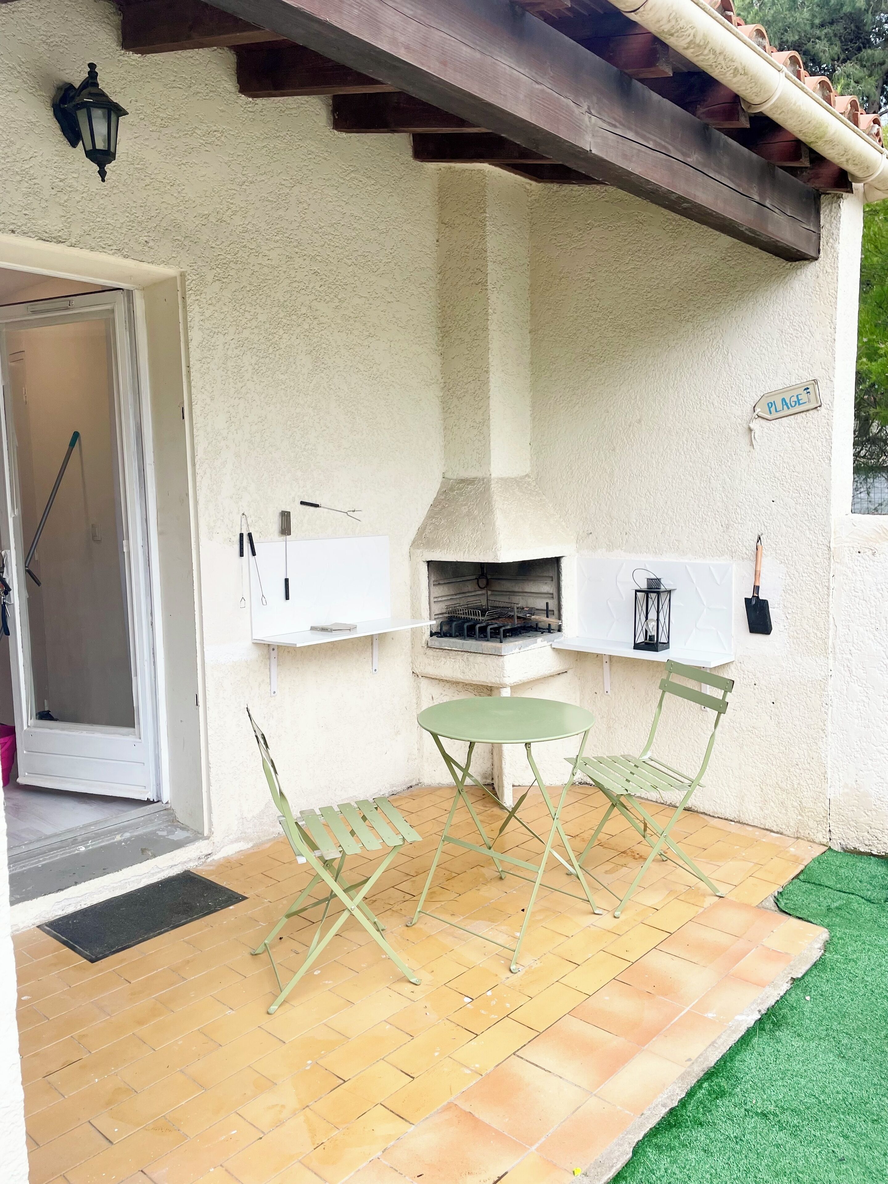 An outdoor patio with a grill, two green chairs, and a white wall with a 'PLAGE' sign.