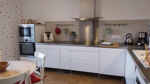 A modern kitchen with white cabinets, a stainless steel range hood, a coffee station, and a dining area with a table and chairs. - Gîte "Chez Tété (Meursault)