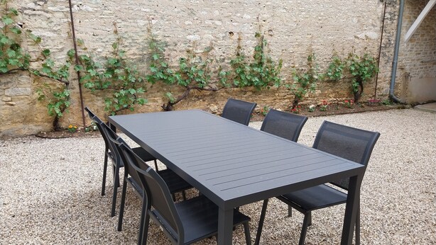 A rectangular outdoor table with four chairs, set on gravel, against a stone wall with climbing plants. - Gîte "Chez Tété (Meursault)