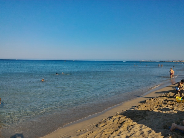 A beach with clear water, people swimming and walking along the shore, and boats in the distance.