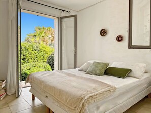 Un dormitorio con cama, almohadas, vistas a la vegetación por una puerta abierta y un reloj de pared.