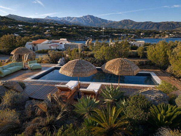 Una piscina con sillones de descanso y sombrillas de paja, rodeada de vegetación y con vistas a un paisaje montañoso.