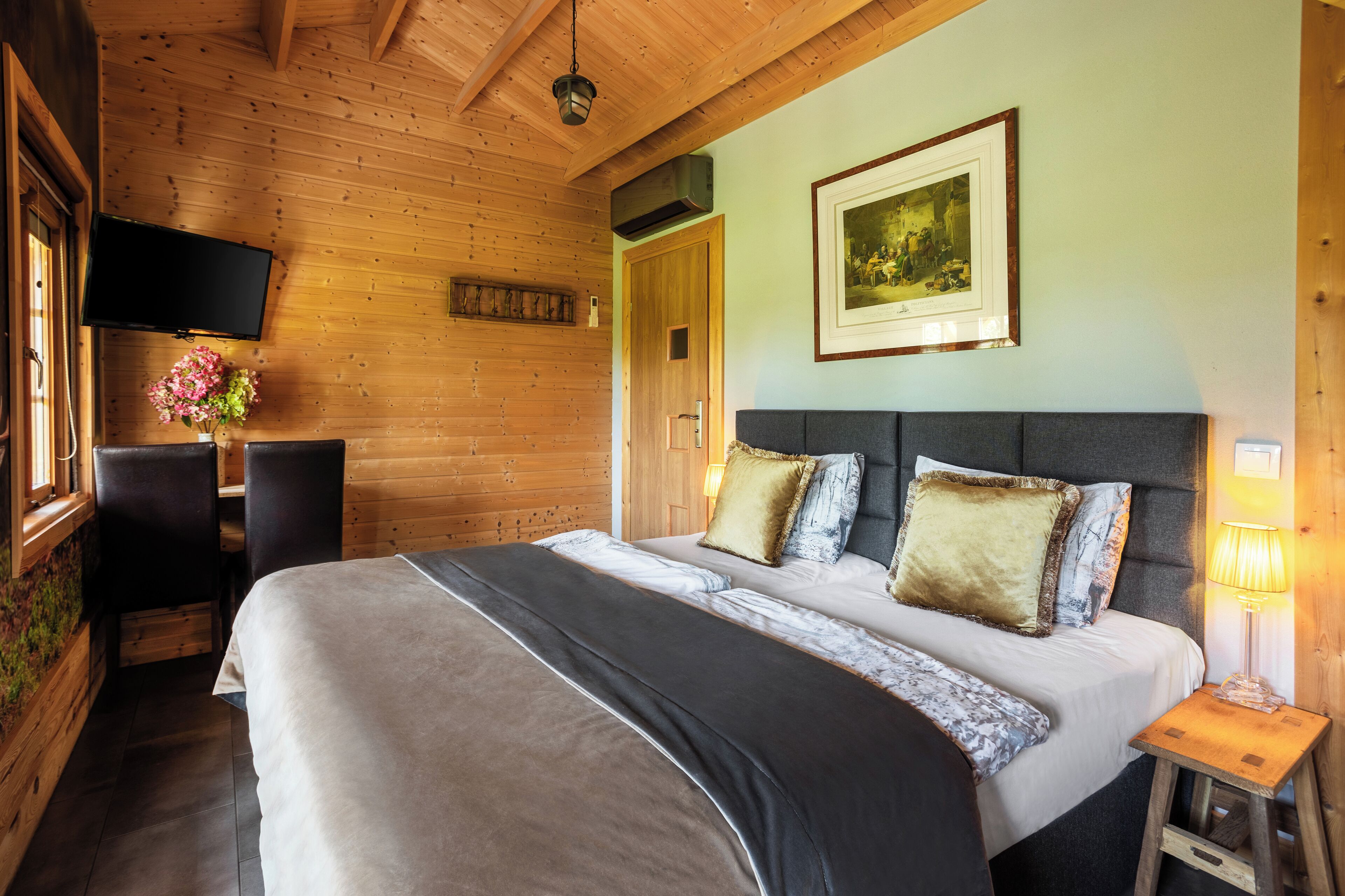 A wooden cabin-style room with a bed, a TV mounted on the wall, a small table with a lamp, and a framed picture above the bed.