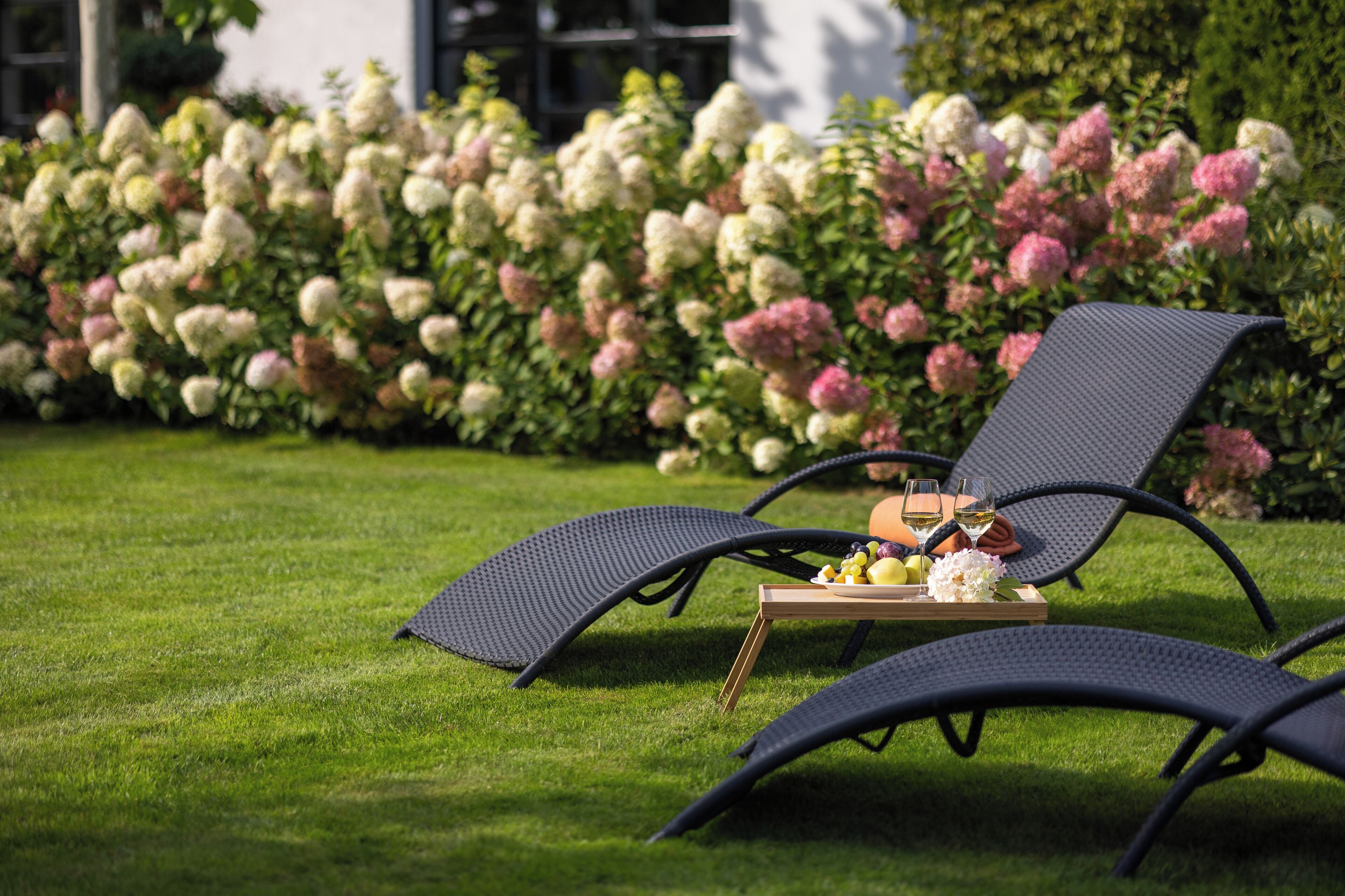 Two outdoor lounge chairs with a tray table and a bottle of wine, set on a well-manicured lawn with a backdrop of flowering bushes.