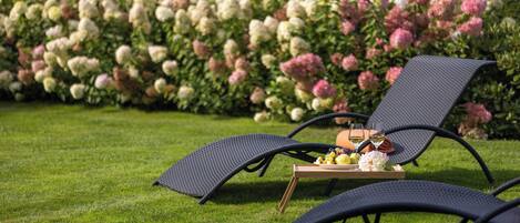 Two outdoor lounge chairs with a tray table and a bottle of wine, set on a well-manicured lawn with a backdrop of flowering bushes.