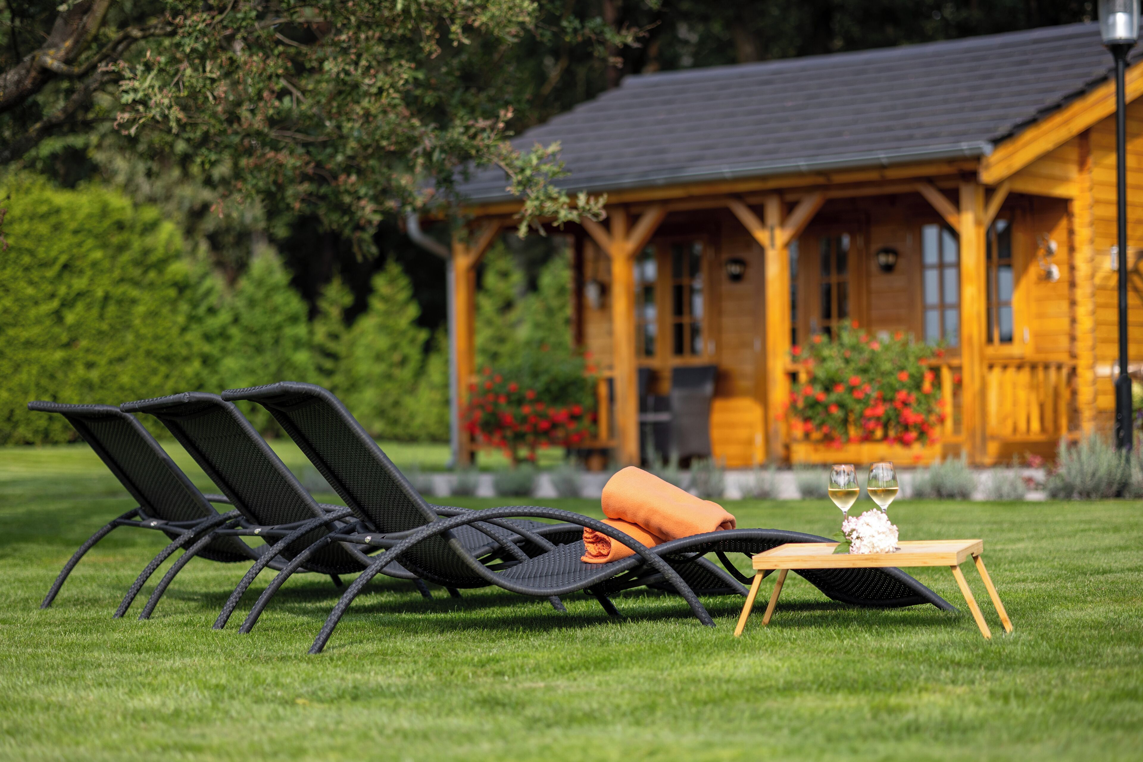 Una cabaña de madera con un balcón, una silla de jardín, una mesa con cojín y dos copas de vino.