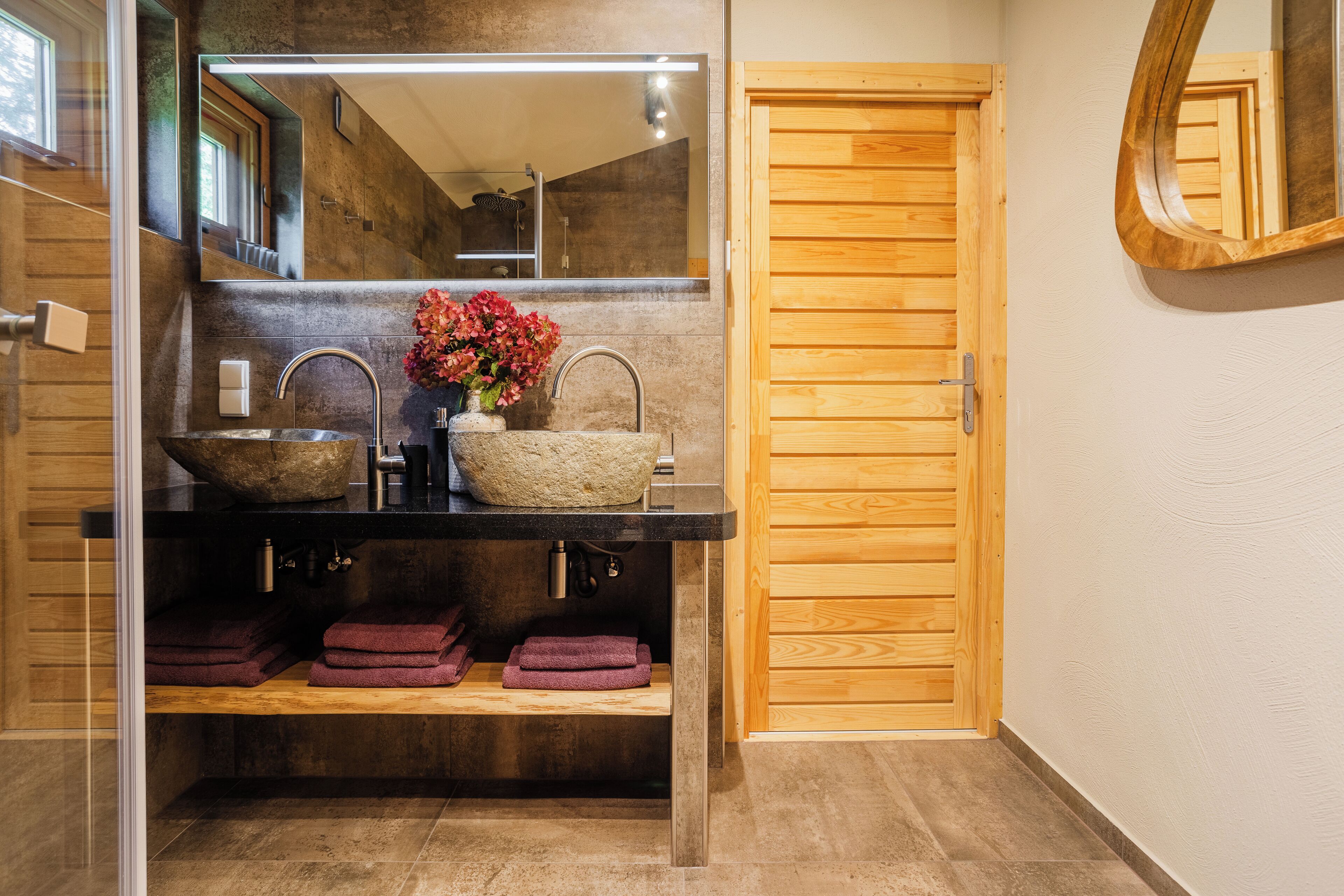 Une salle de bain avec un grand miroir, deux lavabos, une porte en bois et une étagère avec des serviettes.