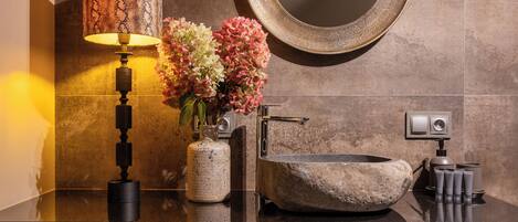 Un baño con lavamanos de piedra, un espejo redondo, un florero con flores y una lámpara.