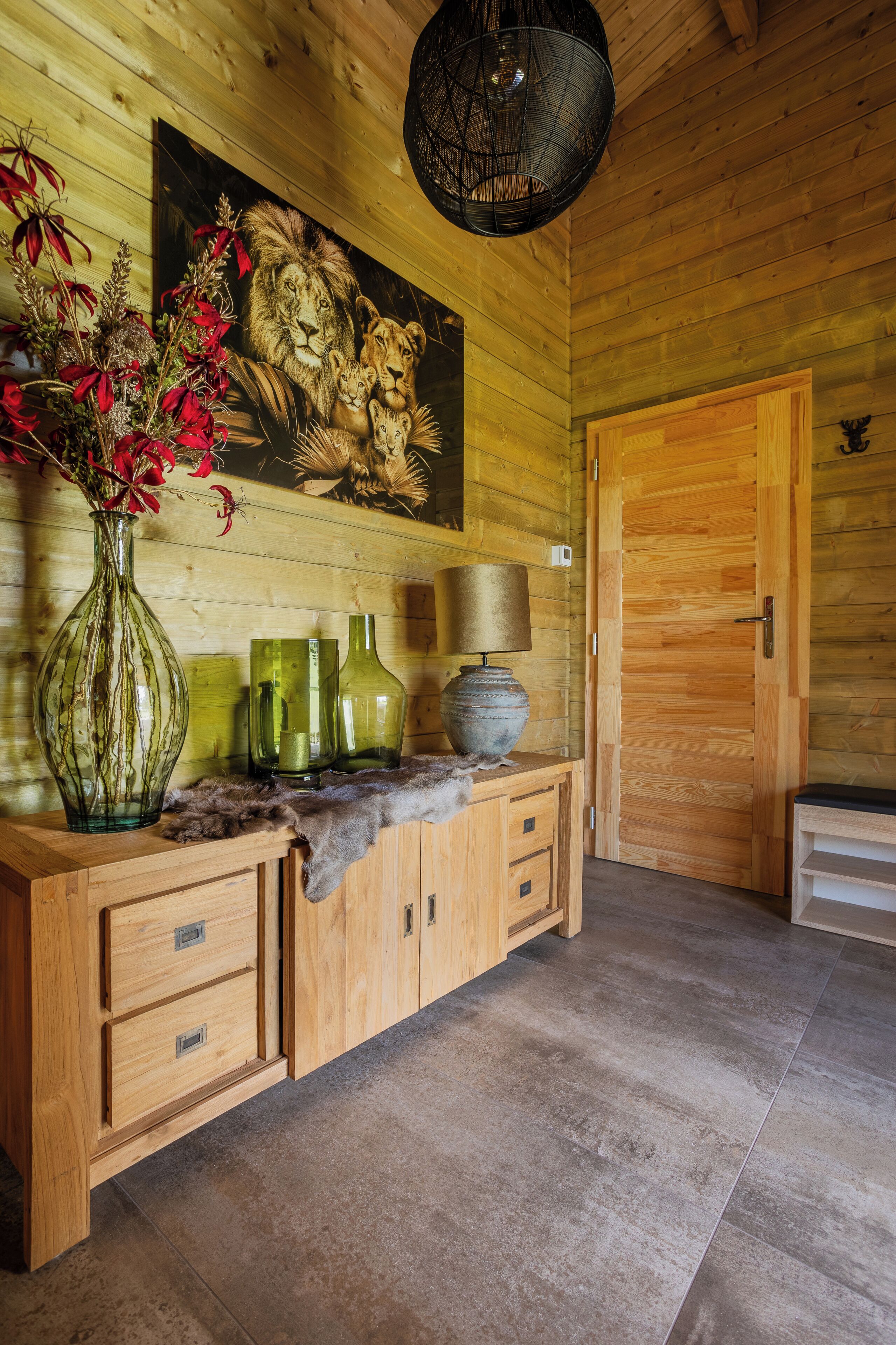 Un intérieur en bois avec un meuble en bois, un vase rempli de fleurs rouges, une lampe et un tableau représentant des lions.