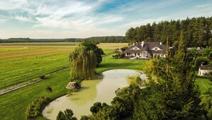 Una casa grande con un estanque, rodeada de campos verdes y árboles.
