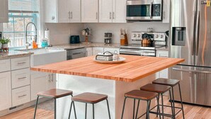 Une cuisine moderne avec des meubles blancs, des appareils en acier inoxydable, un îlot central en bois doté de tabourets et une fenêtre avec des stores.