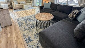 Un salon spacieux avec un canapé gris foncé, une table basse ronde et un tapis à motifs. On y trouve un ventilateur de plafond et une grande fenêtre avec des rideaux.